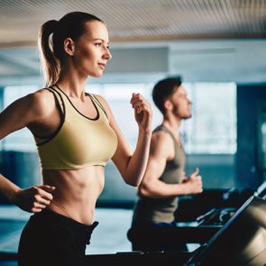 Pretty woman running on treadmill with fit young man on background