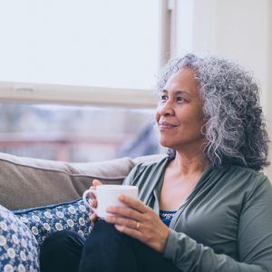 woman sitting on couch at home while holding a cup of coffee and looking out the window