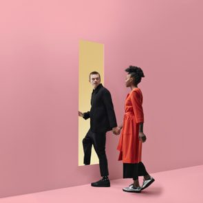 Woman & man holding hands, approaching rectangular opening in coloured wall